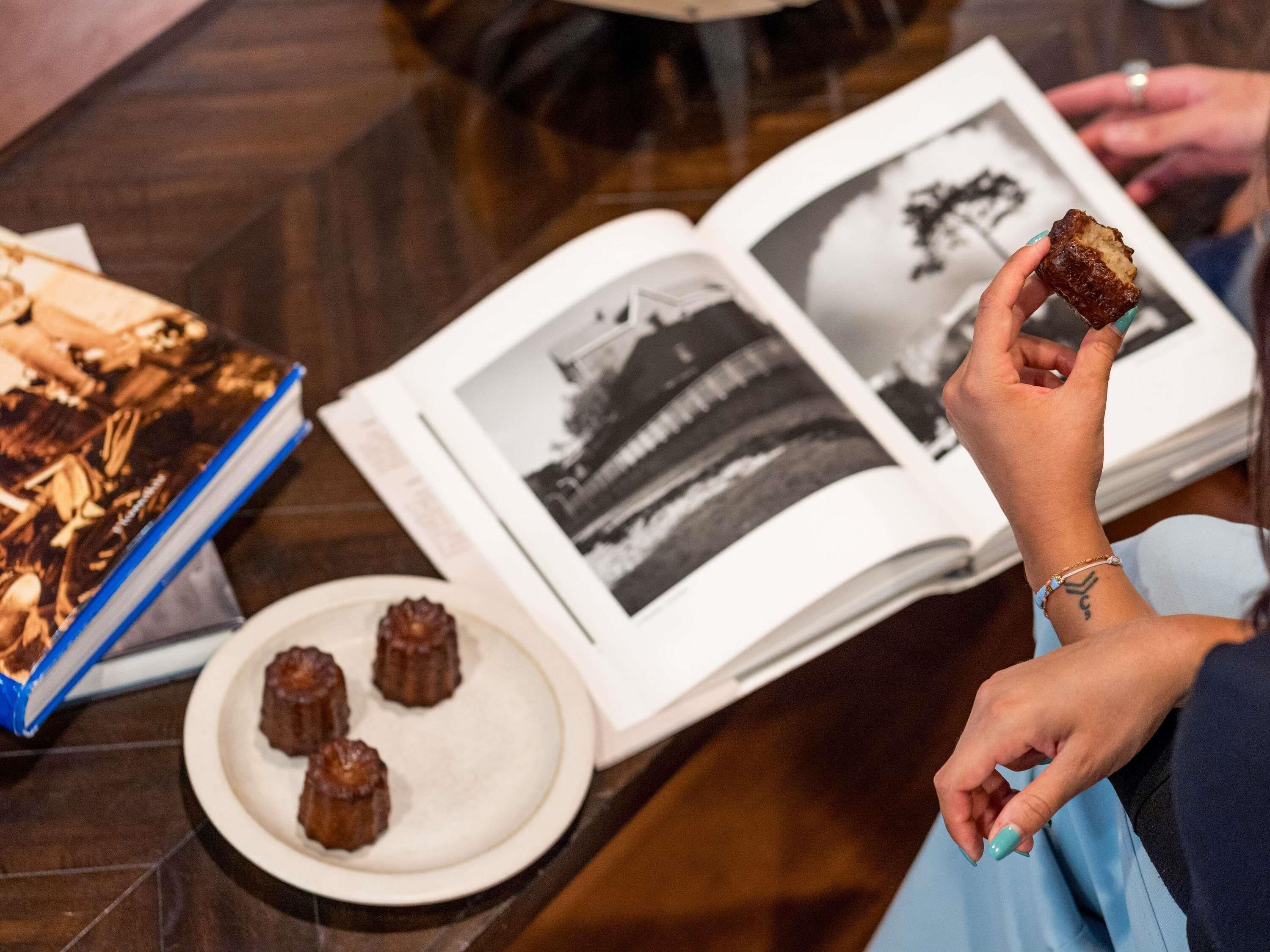 A person holding a pastry, a plate with three pastries, and an open book with a black and white photo on a table.