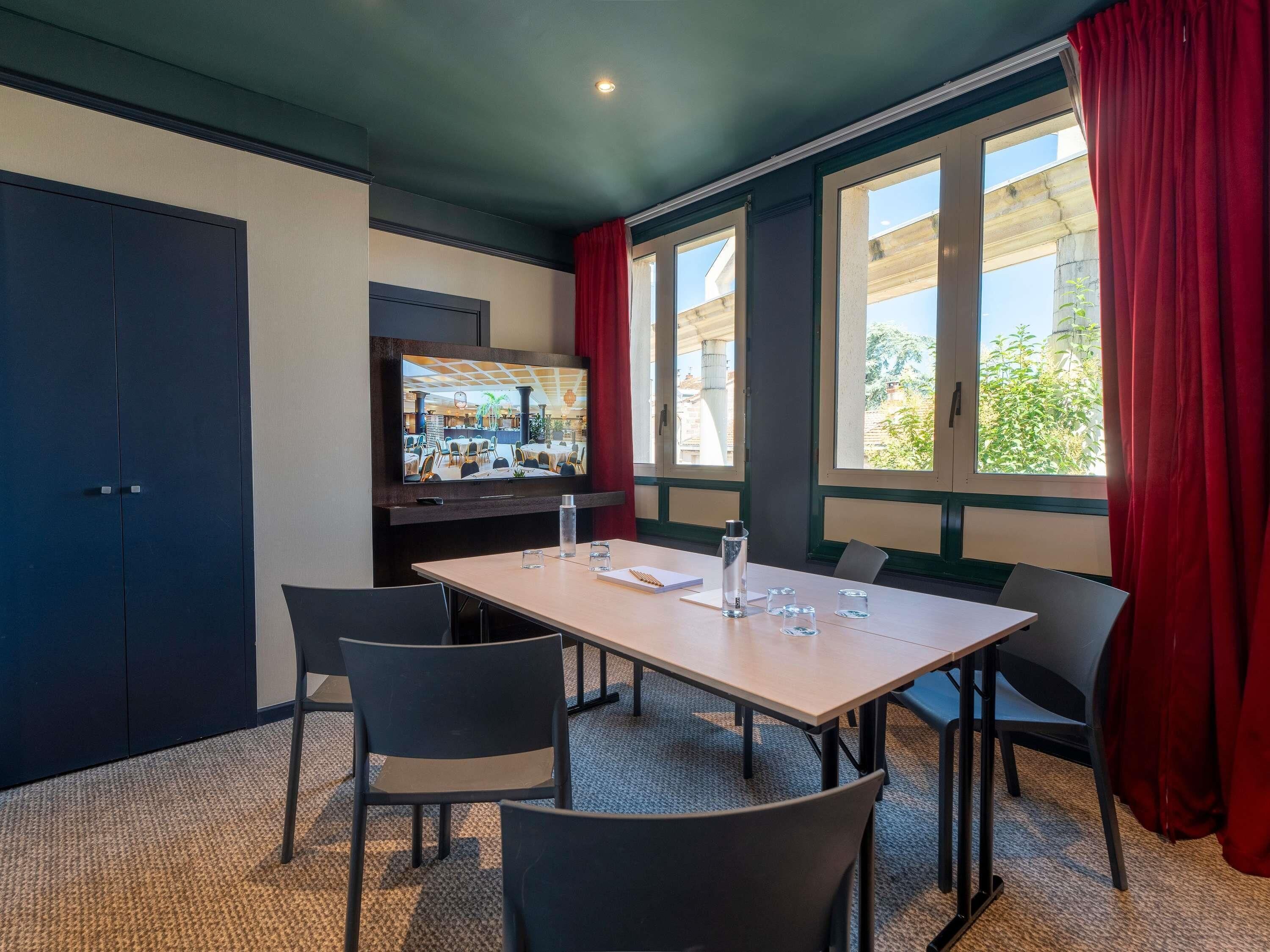 A conference room with a large table, chairs, a television screen, and windows with curtains.