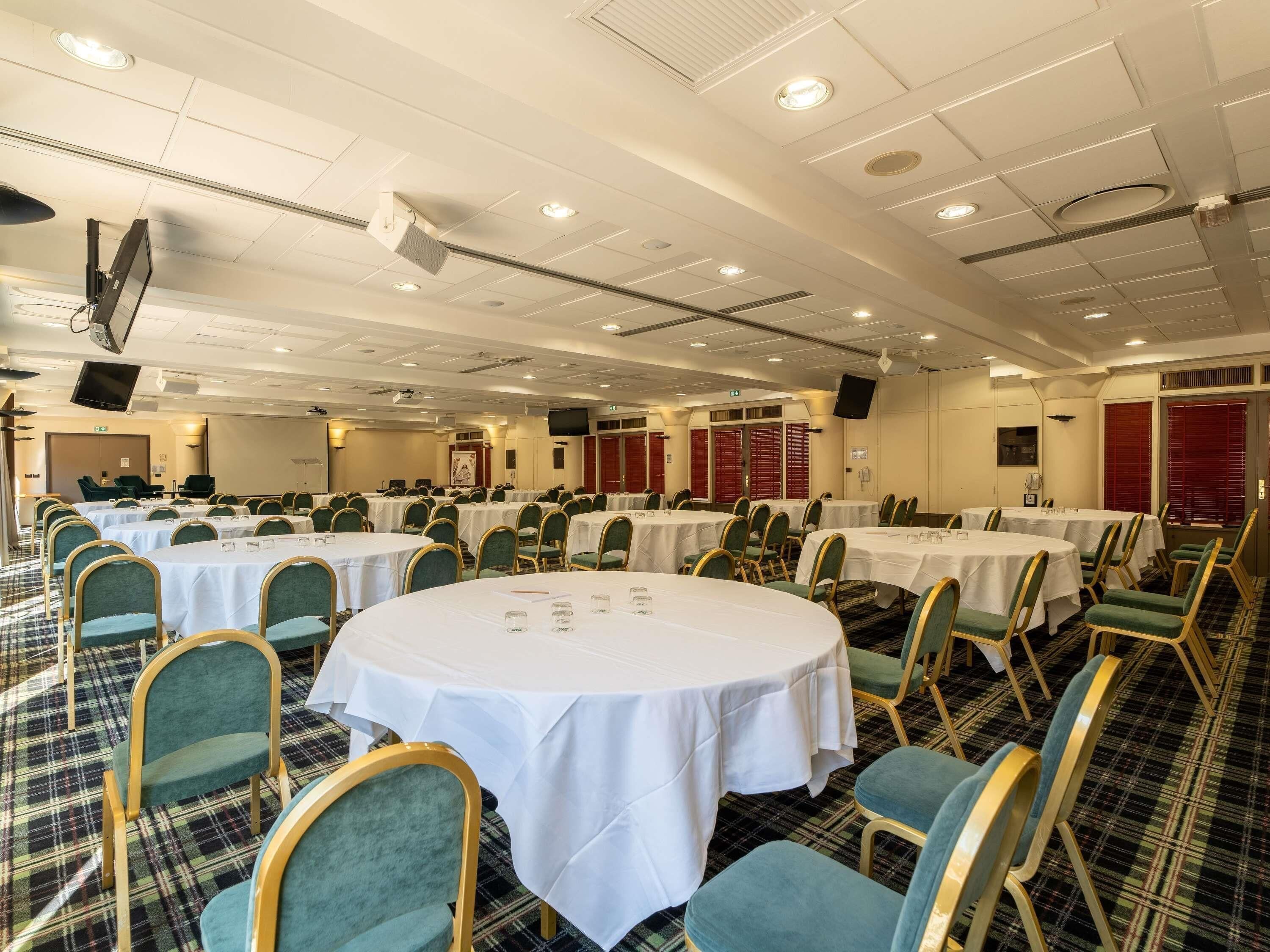 Un salón de conferencias con varias mesas y sillas, equipado con un proyector y una pantalla.