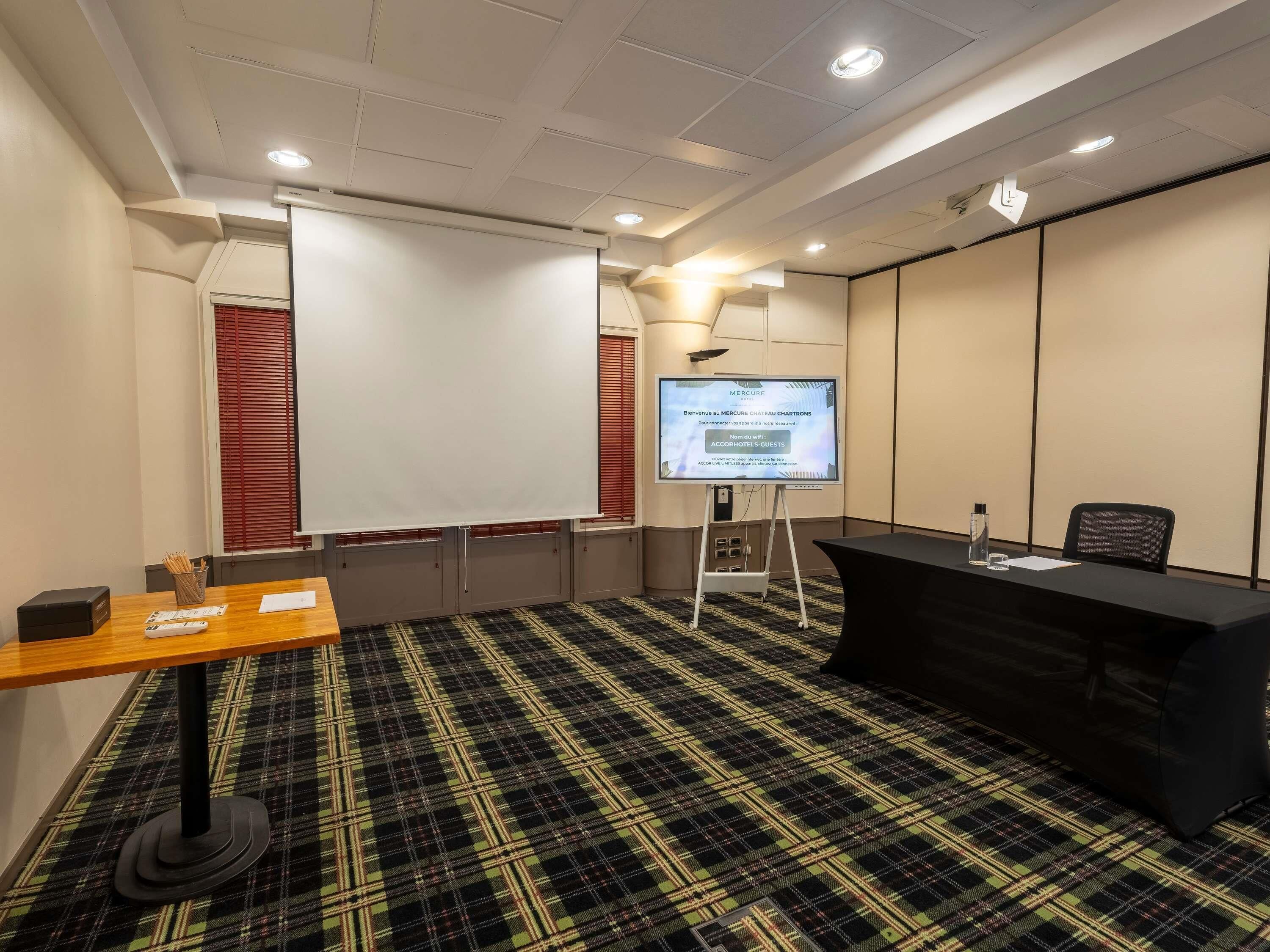 A conference room with a large screen, a wooden table, a black conference table, and a plaid carpet.