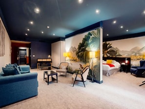 Una habitación de hotel moderna con sofá, sillones, una mesa de comedor, una cama y un mural grande en la pared.