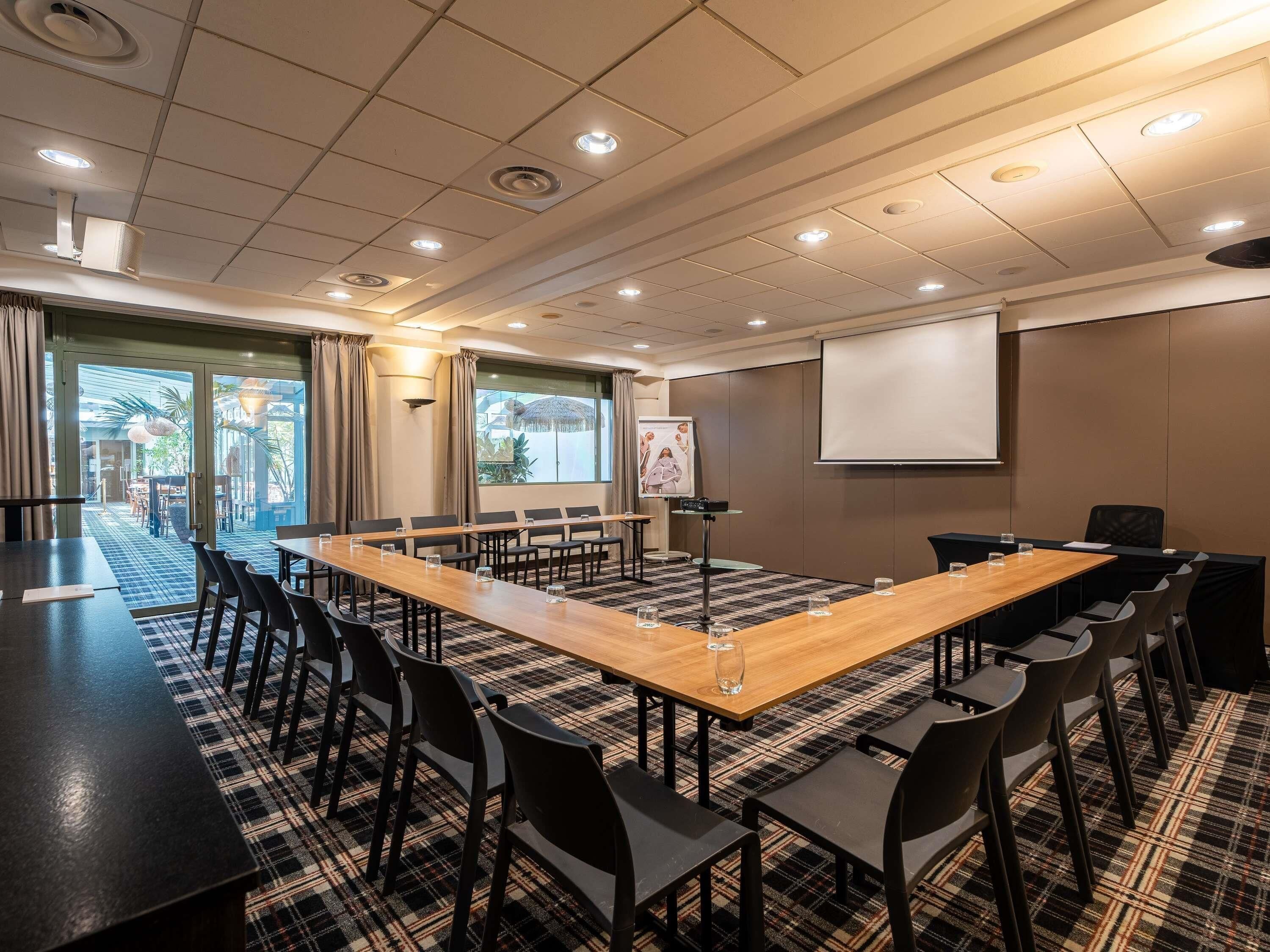 Ein Konferenzraum mit langem Tisch, Stühlen, einer Projektionsleinwand und einem großen Fenster.