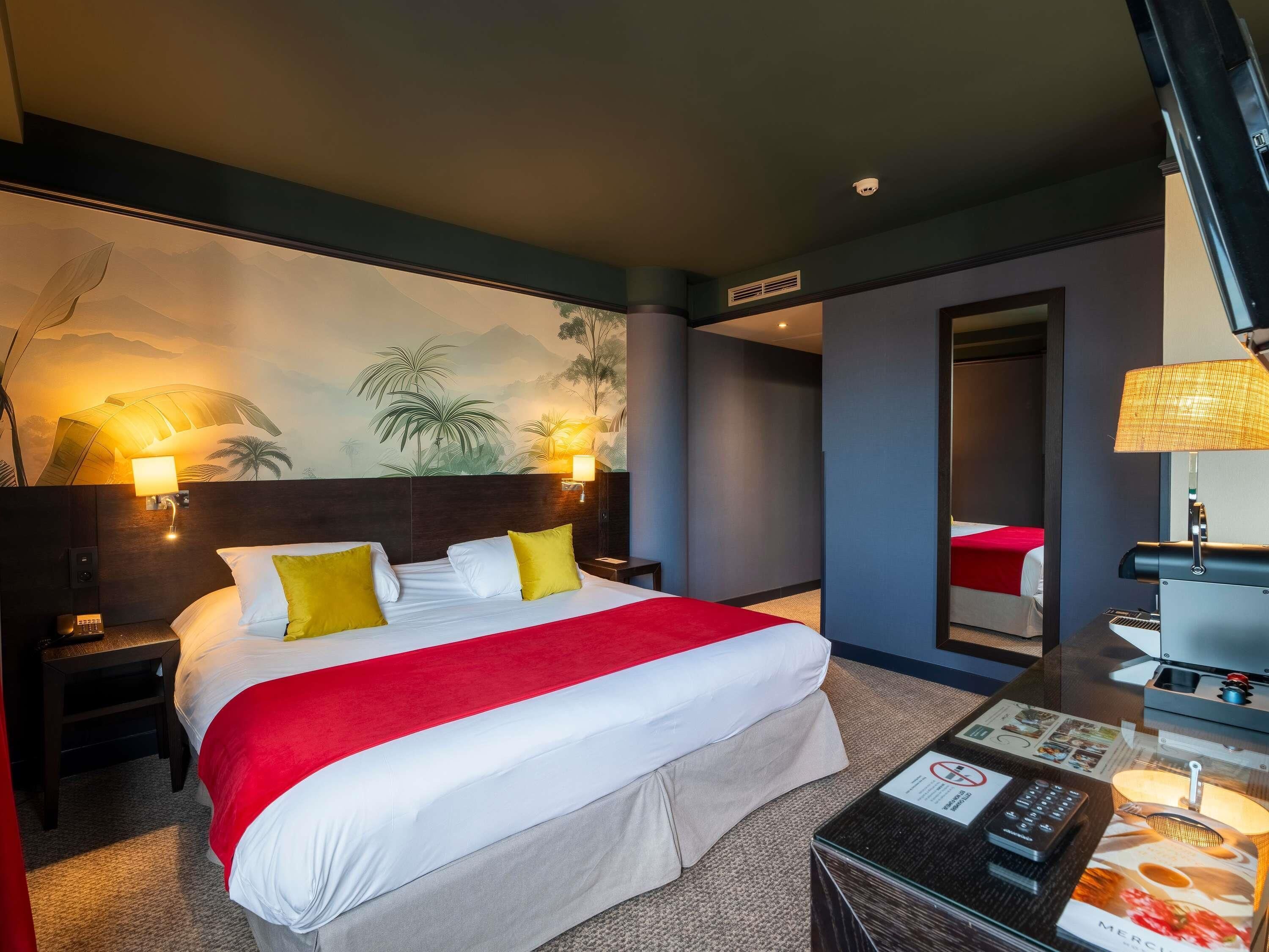 Une chambre d’hôtel avec un grand lit, deux tables de chevet, un bureau avec une lampe, un climatiseur mural et une fresque représentant un paysage tropical.