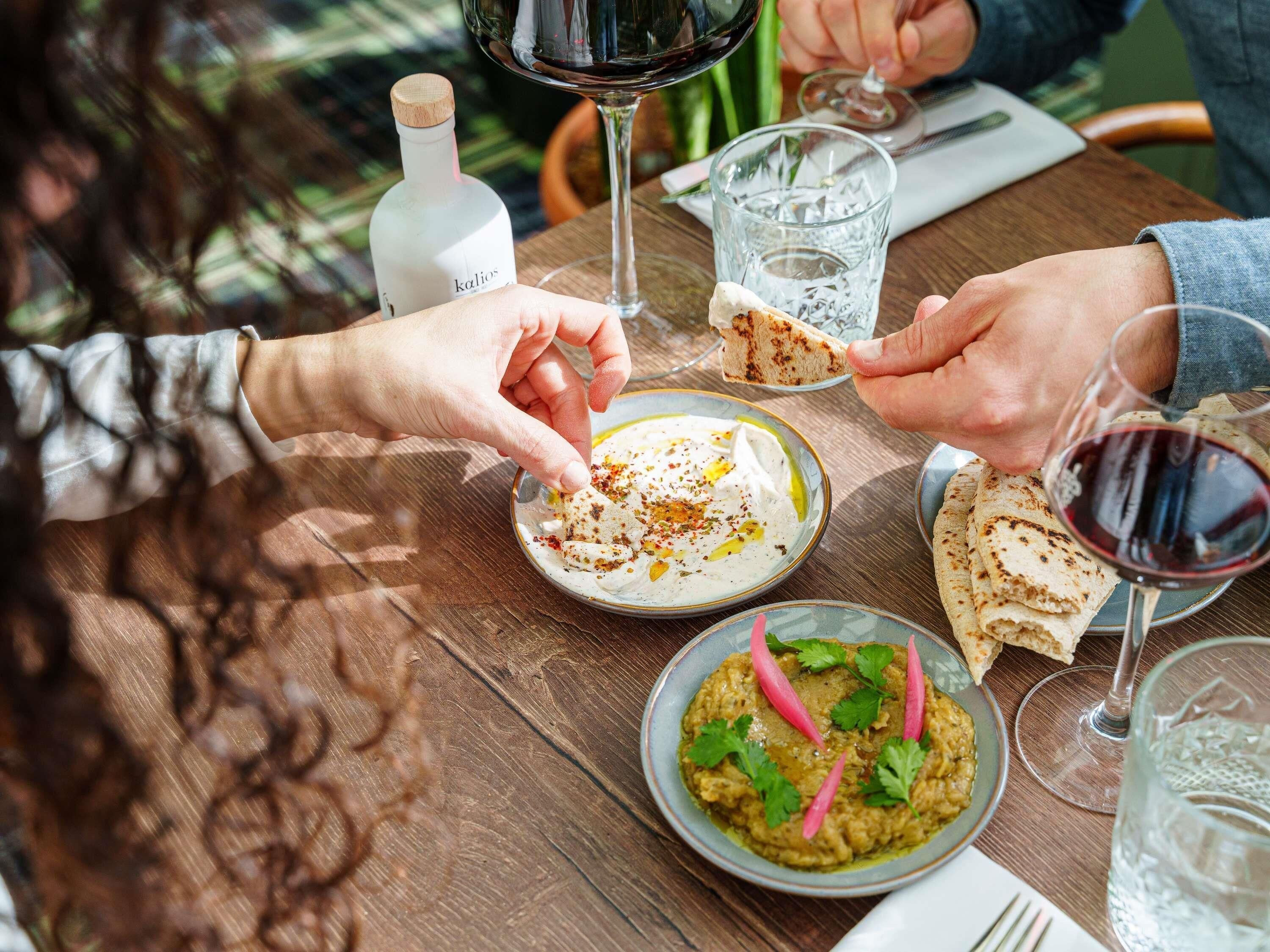 Ein Holztisch mit einer Schüssel weißer Dip, garniert mit Kräutern, einem Teller Fladenbrot und einer Flasche Flüssigkeit.