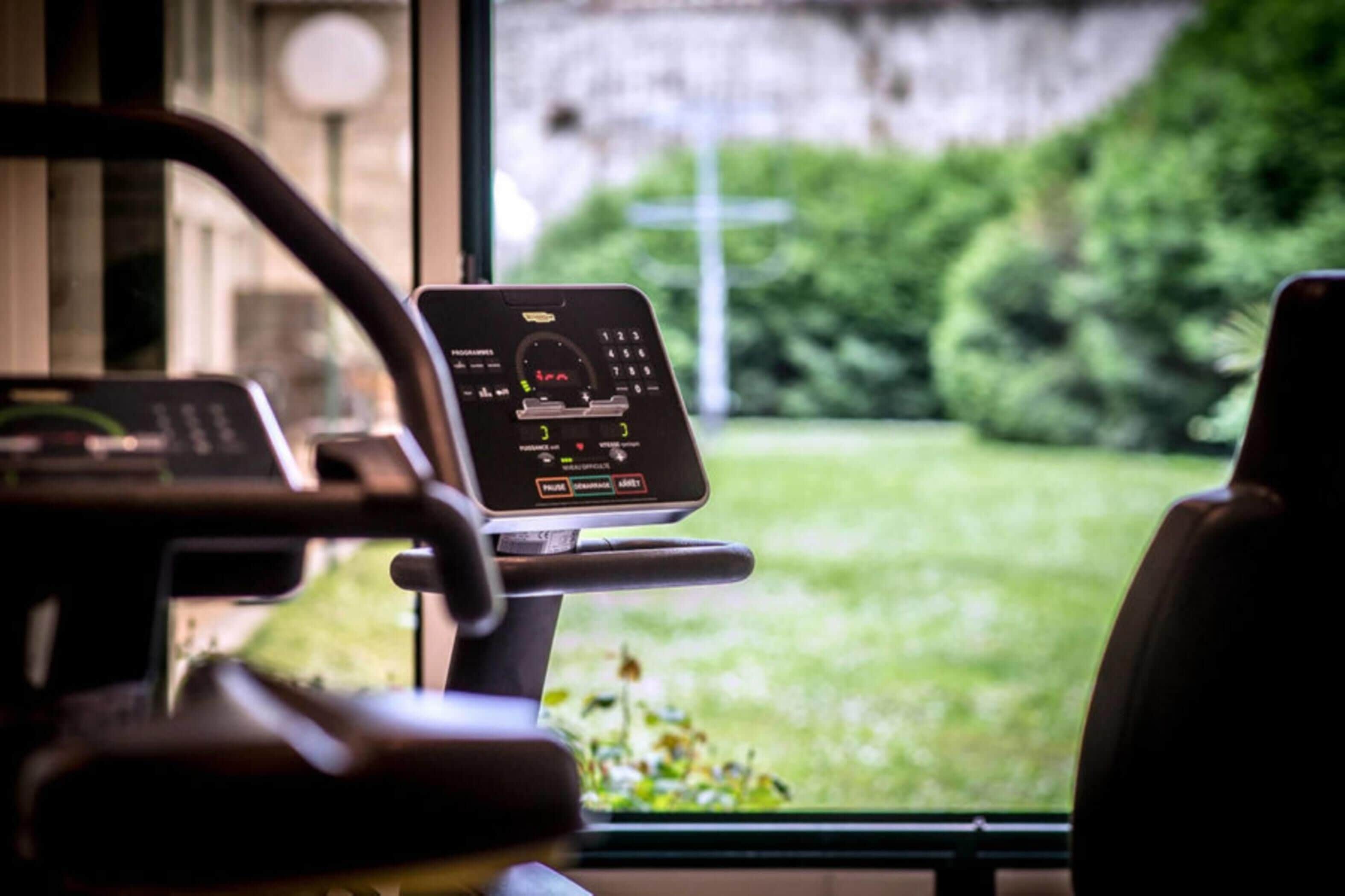 A stationary exercise bike with a digital display, positioned in front of a window with a view of a green landscape.