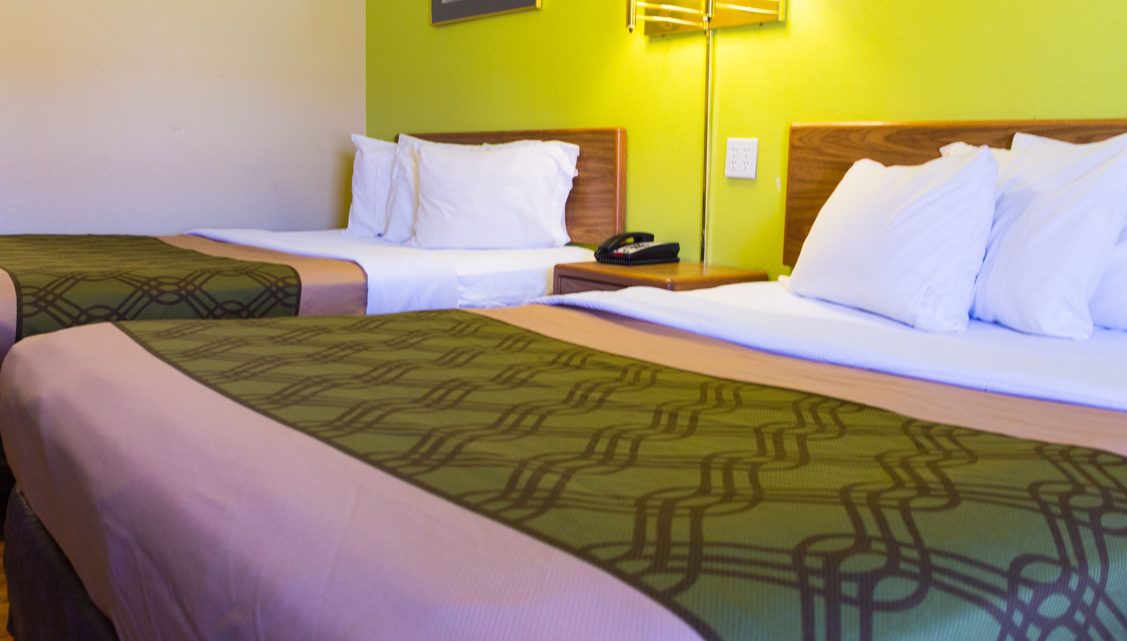 A hotel room with two beds, each with white linens and a patterned green bedspread. There is a bedside table with a telephone and a lamp on one side.