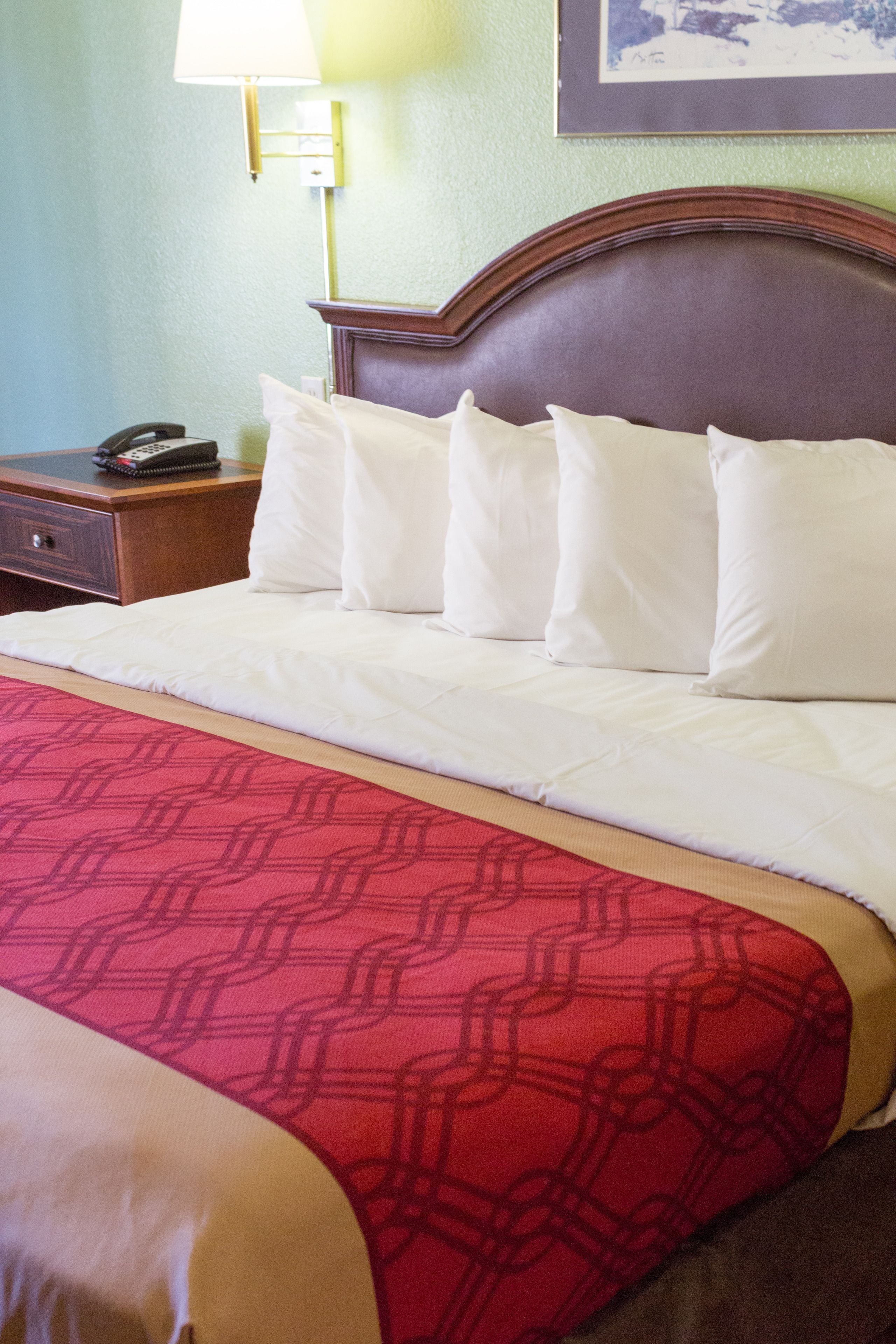 Ein Hotelzimmer mit einem ordentlich bezogenem Bett, einer roten, gemusterten Tagesdecke, einem Nachttisch aus Holz mit Telefon und einer Wandlampe.
