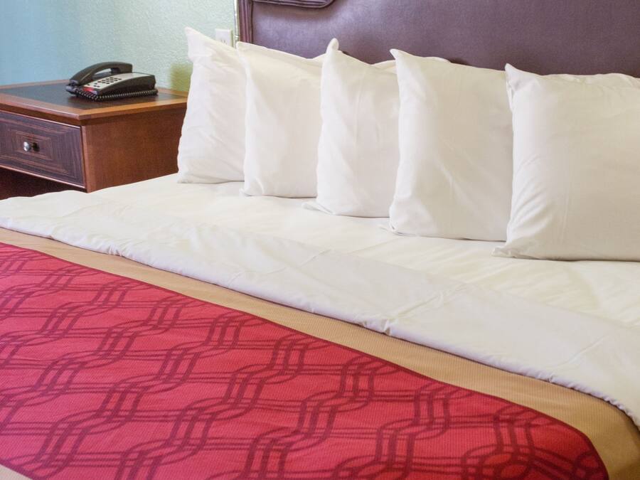 Ein Hotelzimmer mit einem ordentlich bezogenem Bett, einer roten, gemusterten Tagesdecke, einem Nachttisch aus Holz mit Telefon und einer Wandlampe.
