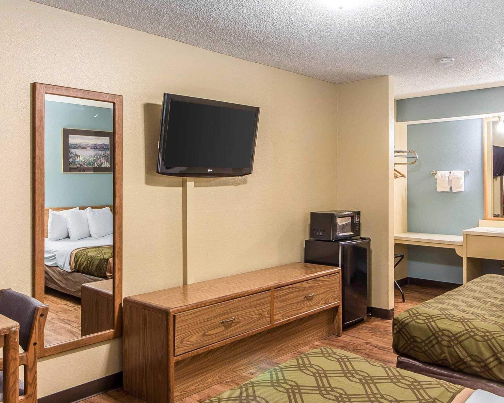 A hotel room with a bed, a wooden dresser, a television mounted on the wall, a microwave, and a small refrigerator.