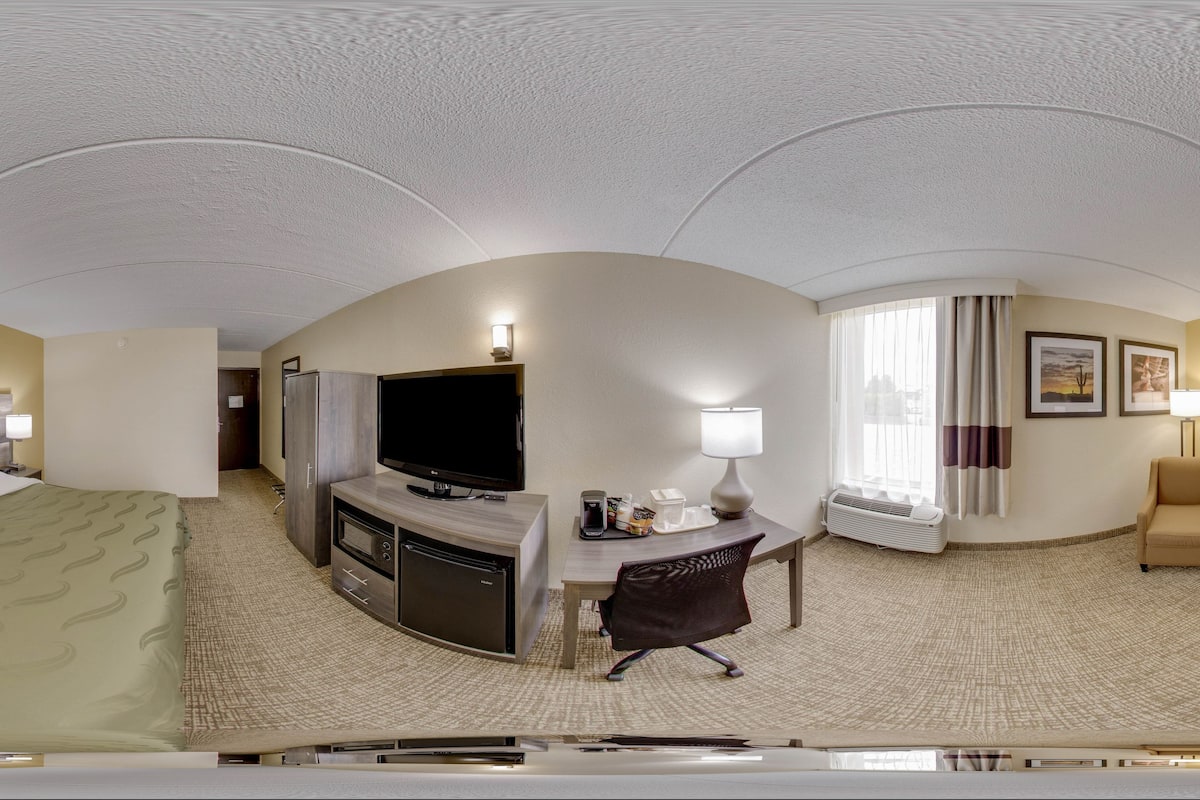 A hotel room with a bed, a TV, a desk with a lamp, a chair, and a window with curtains.
