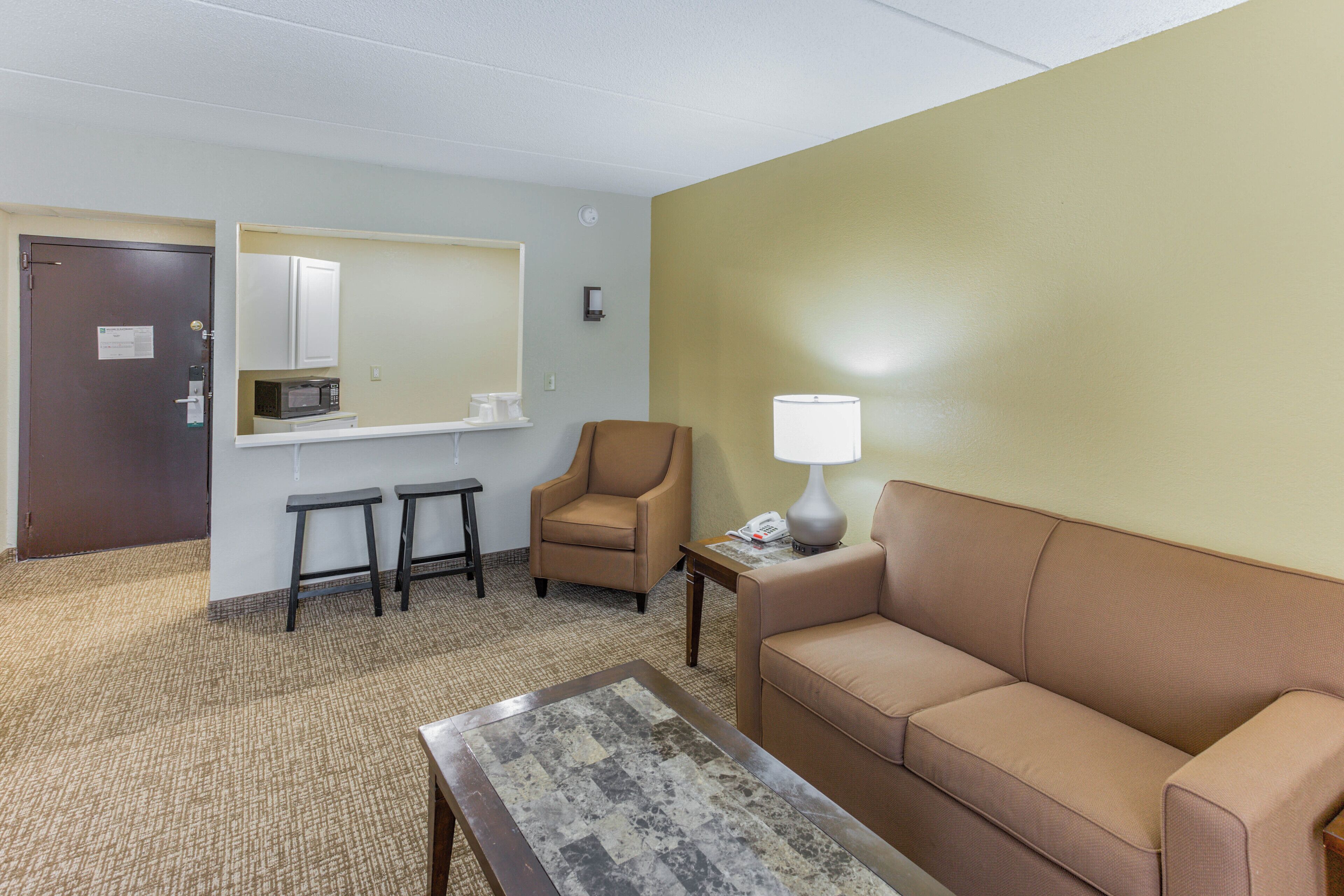 A hotel room with a brown sofa, a coffee table, a small armchair, a microwave, a sink, and a mirror.