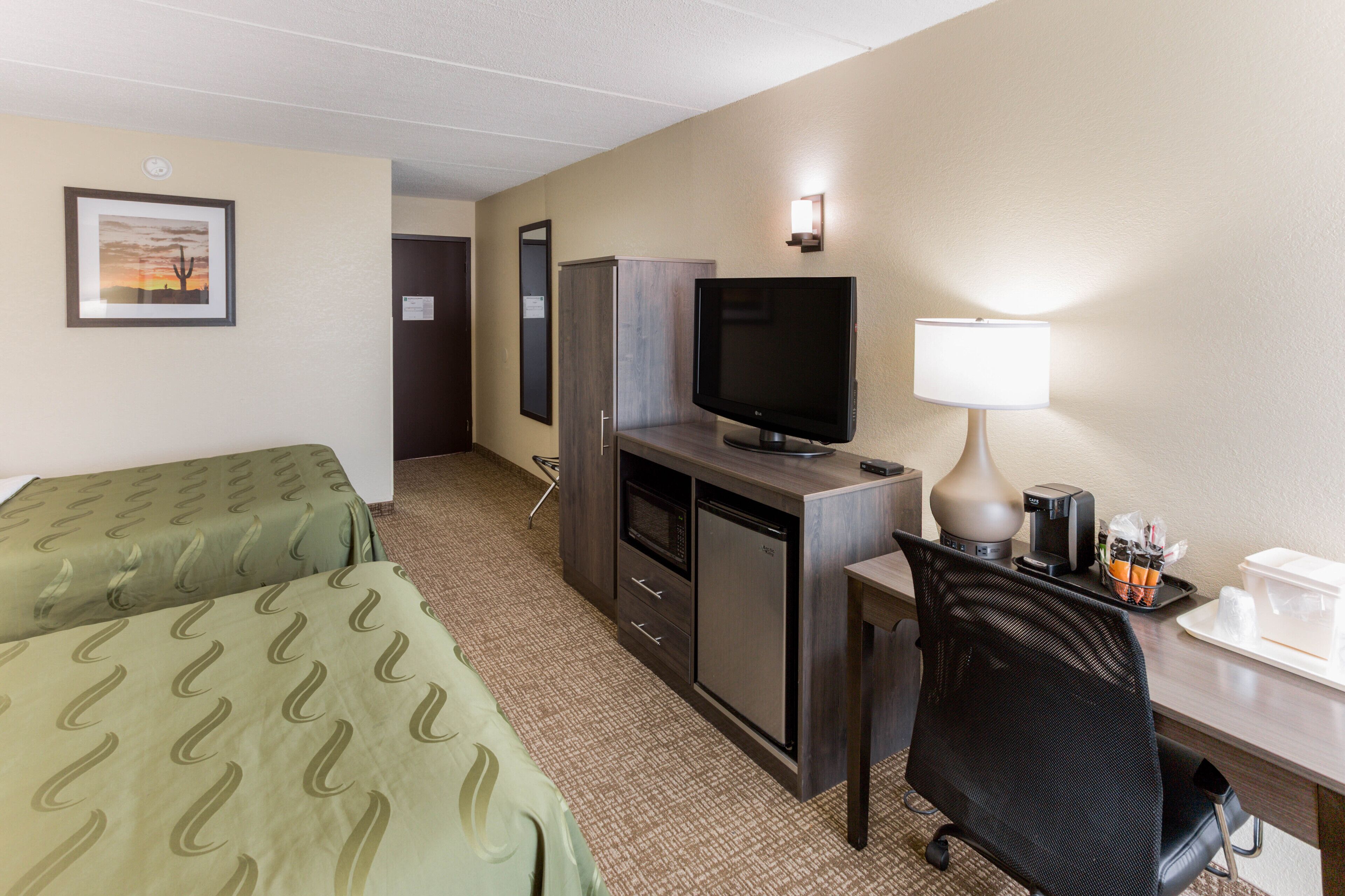 A hotel room with two single beds, a desk with a television, a lamp, and a small refrigerator.