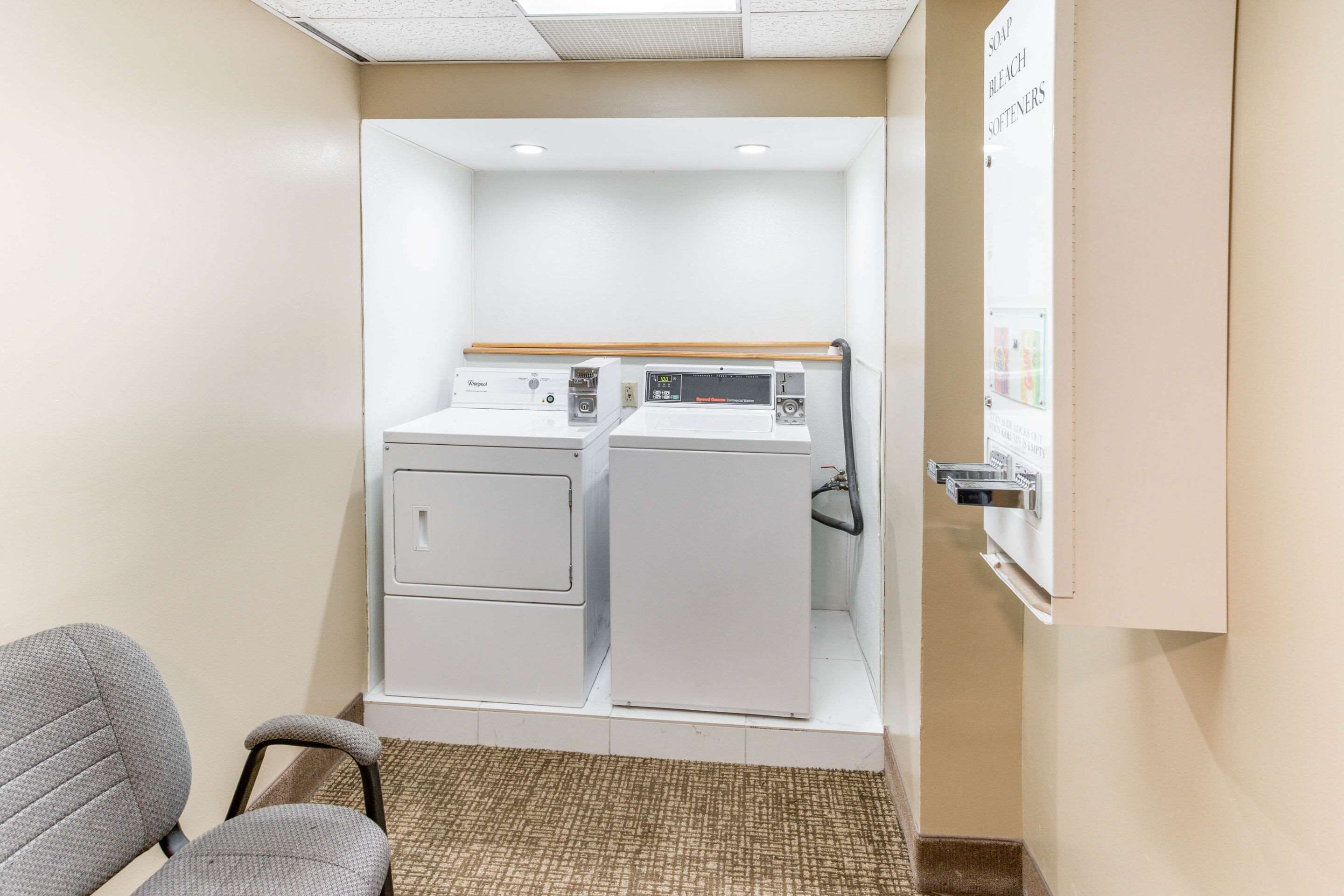 A laundry room with a washing machine and dryer, a small seating area with a chair, and a wall-mounted information board.
