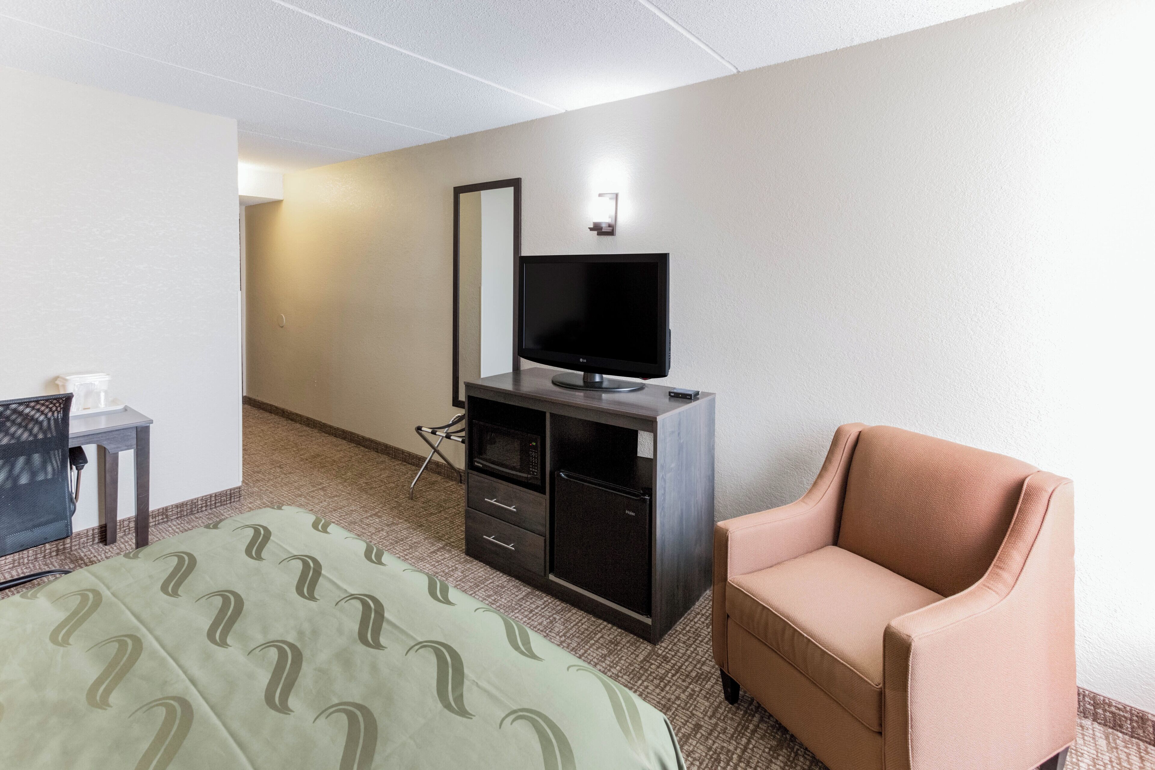 A hotel room with a bed, a television on a stand, a small desk with a chair, and a sofa.