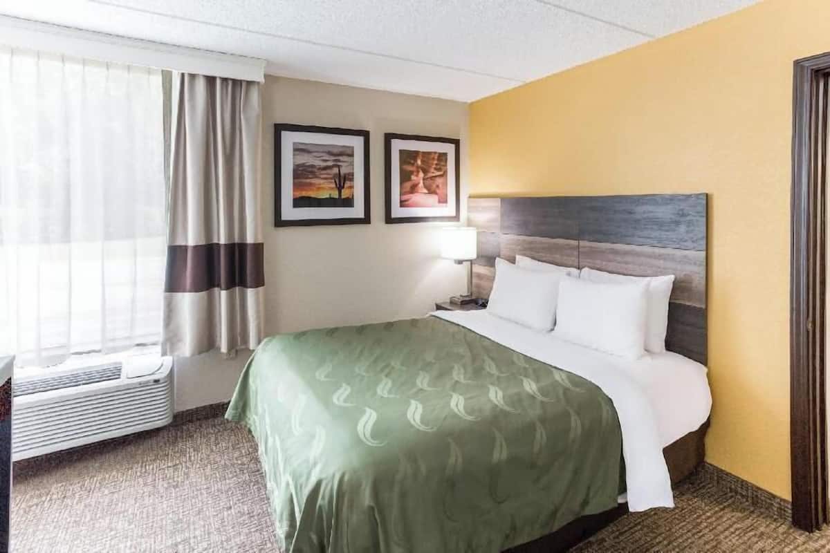 A hotel room with a bed, a window with curtains, a wall-mounted air conditioner, and two framed pictures.