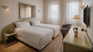 A hotel room with two beds, a desk with a lamp, a chair, a TV, and a potted plant. - NH Ancona (Ancona)