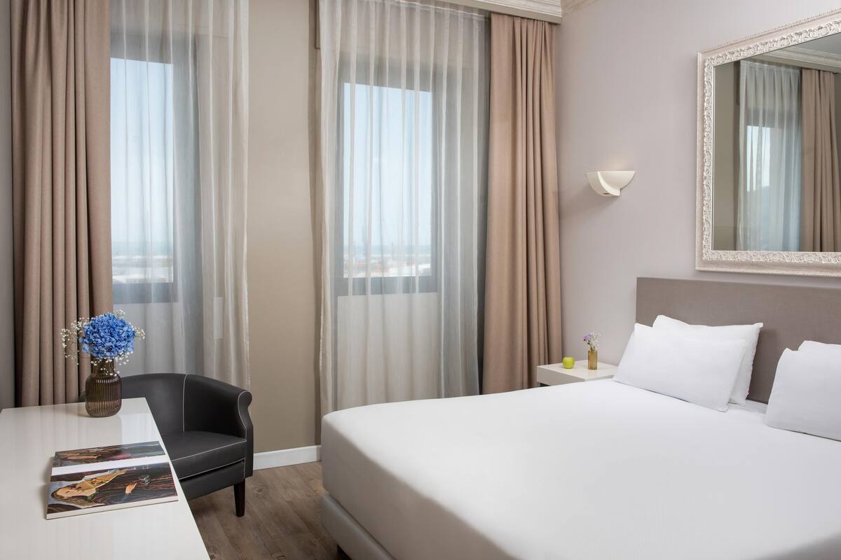 A hotel room with a bed, a chair, a table, and a vase with flowers.