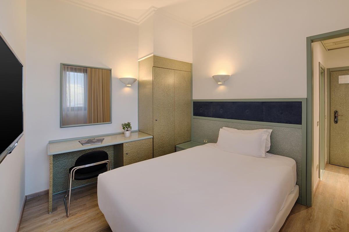 A hotel room with a bed, a desk with a chair, a mirror, and two wall-mounted lights.