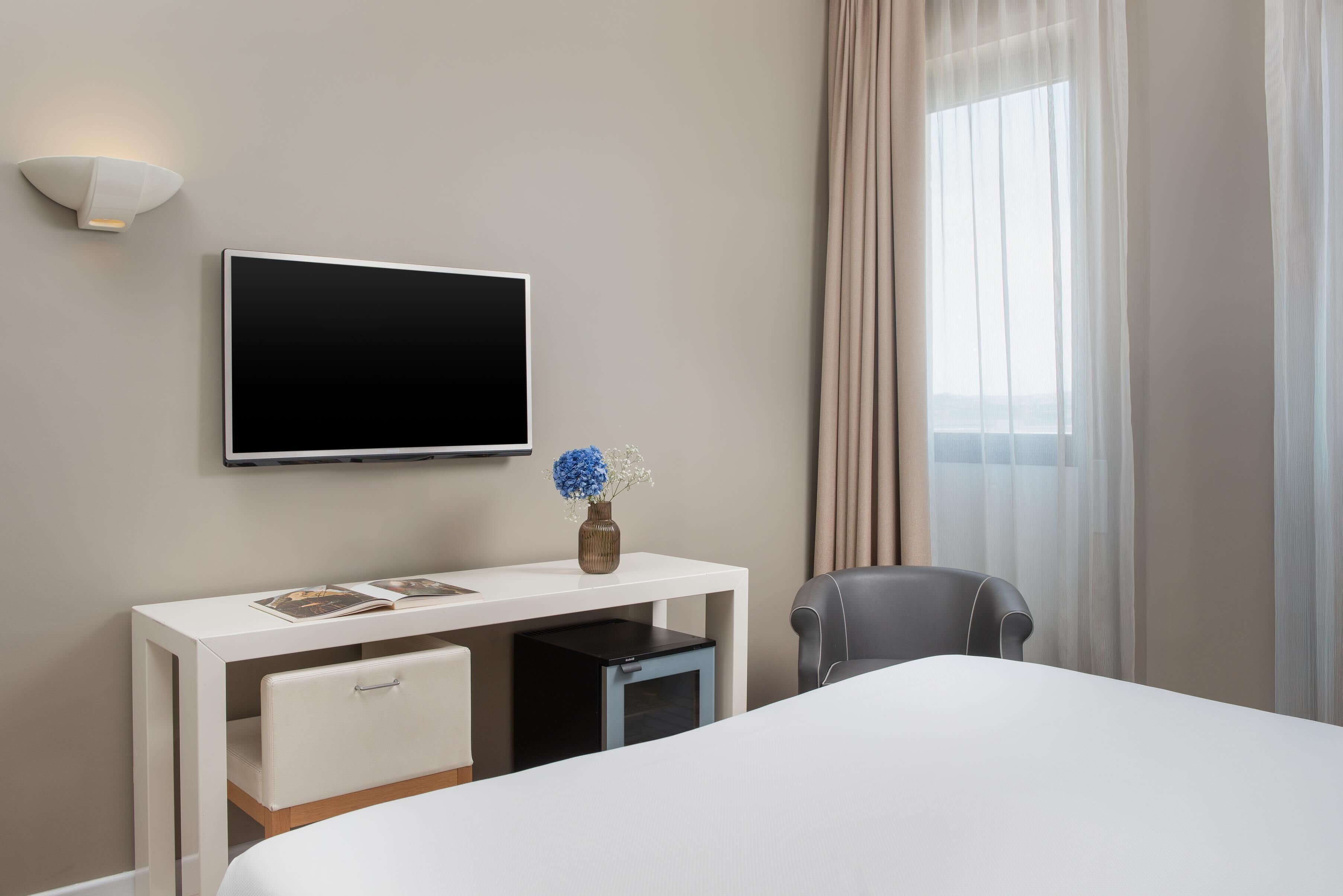 A hotel room with a bed, a desk with a flat-screen TV, a chair, a small cabinet, and a vase with flowers.
