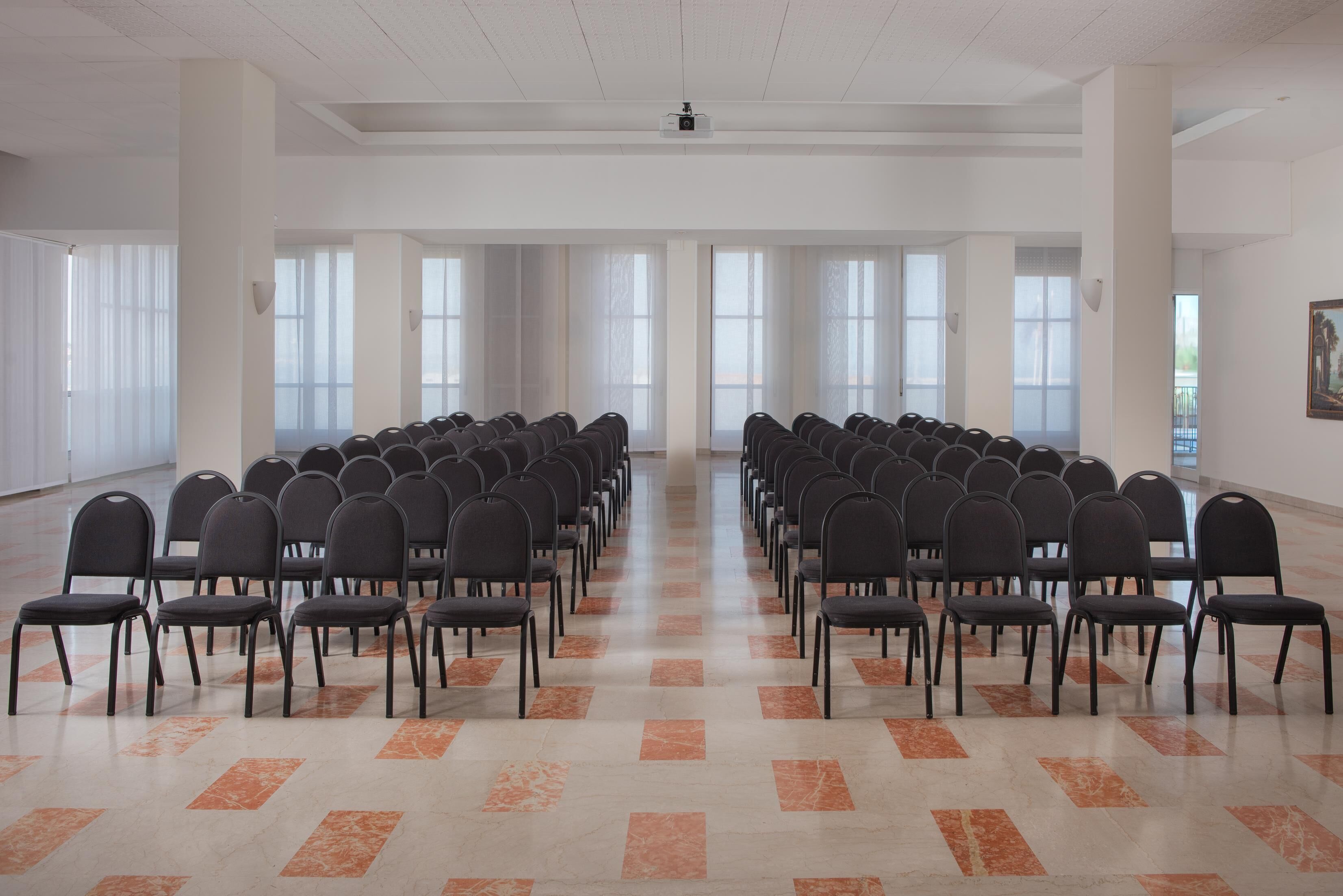 Une salle de conférence avec des rangées de chaises disposées face à un écran de projection, de grandes fenêtres et un sol carrelé.