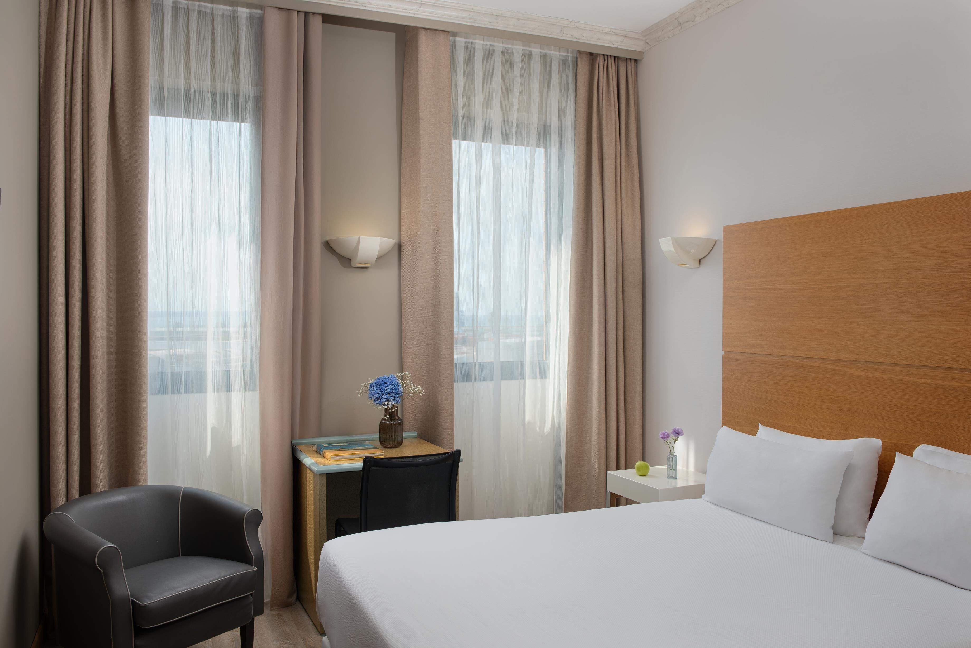 A hotel room with a large bed, a desk with a vase of flowers, a chair, and a view of the city through sheer curtains.