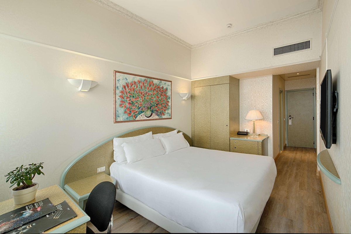 Une chambre d’hôtel avec un grand lit, un bureau agrémenté d’une plante, un tableau au mur, une télévision et une porte donnant sur une autre pièce.