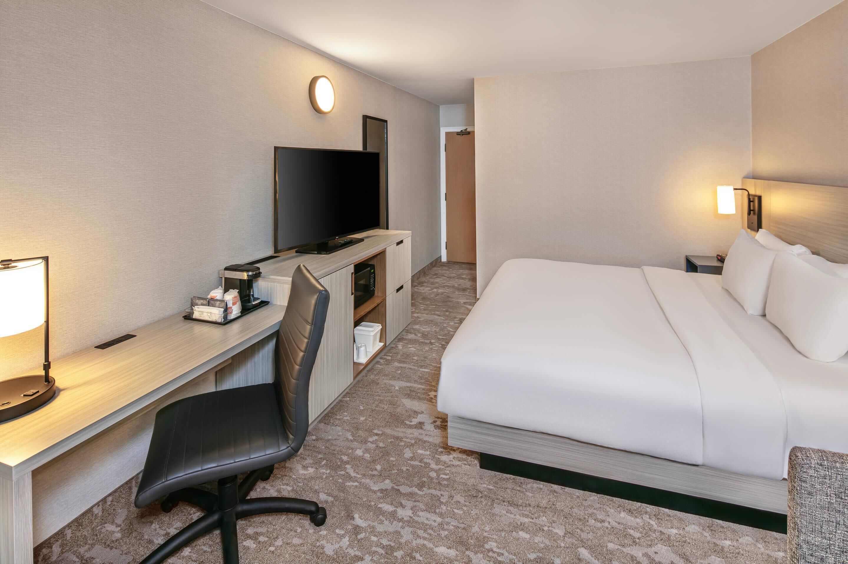 A hotel room with a bed, a desk with a chair, a television, and a nightstand with a lamp.
