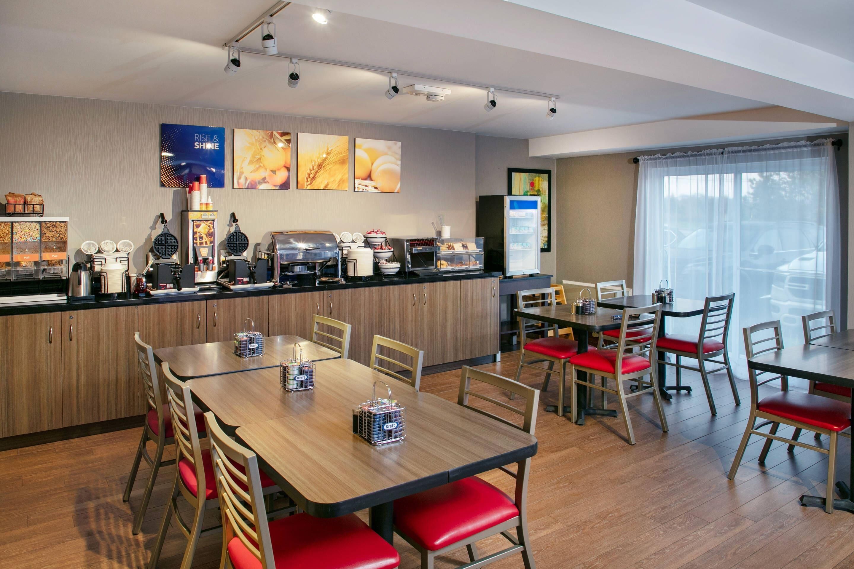 Een moderne hotel eetruimte met houten tafels en rode, beklede stoelen, een buffet met diverse gerechten en een koelkast.