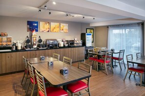 Comedor moderno de hotel con mesas de madera y sillas rojas con cojines, una estación de buffet con distintos alimentos y un refrigerador.