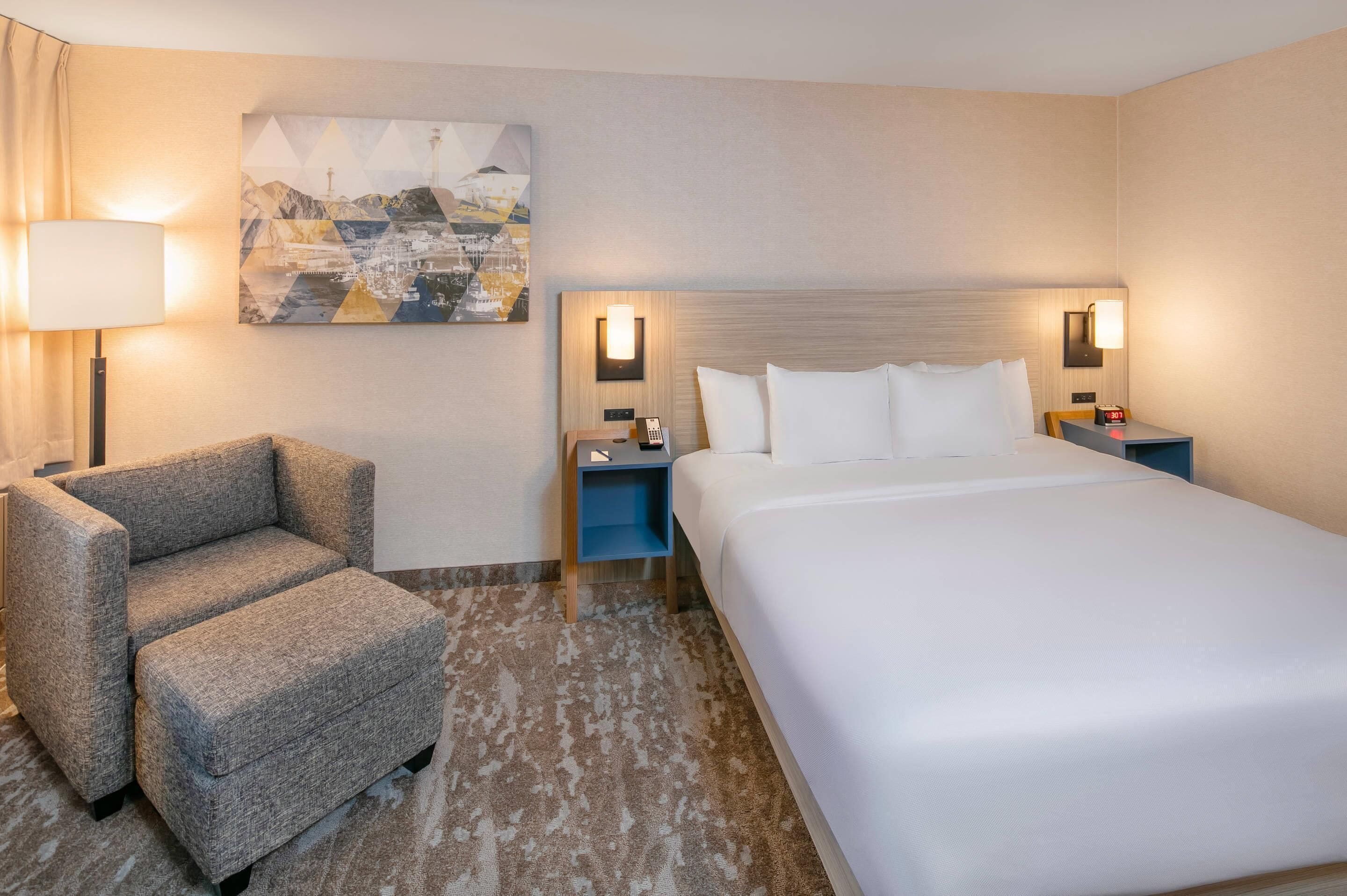 Ein Hotelzimmer mit einem großen Bett, zwei Nachttischen, einem Sessel, einer Lampe und einem Bild an der Wand.