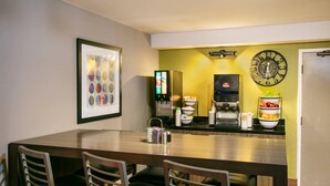 A hotel room with a dining area featuring a wooden table, four chairs, a coffee machine, a clock, and a wall-mounted picture.