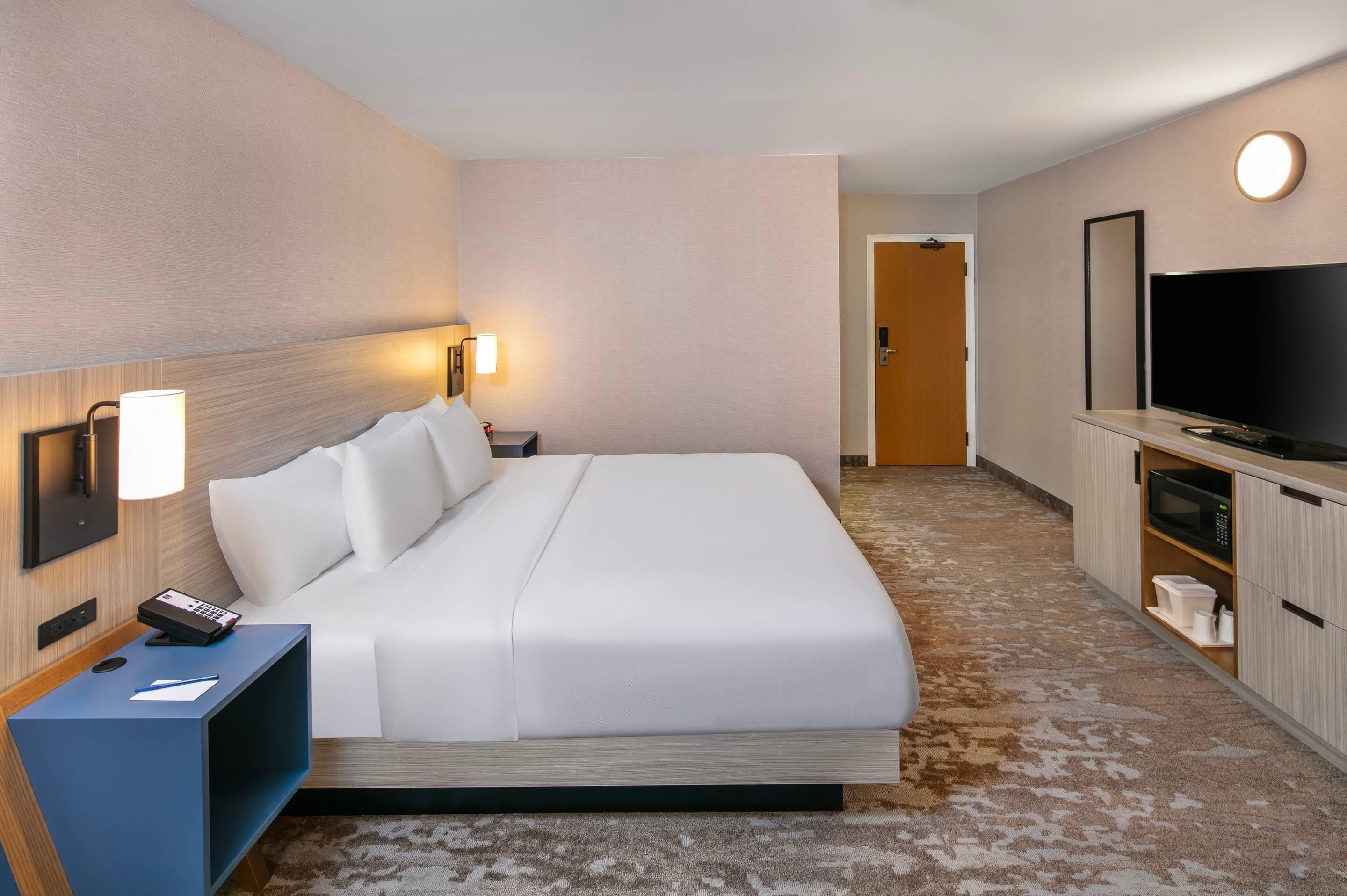 Een hotelkamer met een bed, nachtkastje, lamp, televisie en een klein keukenhoekje.