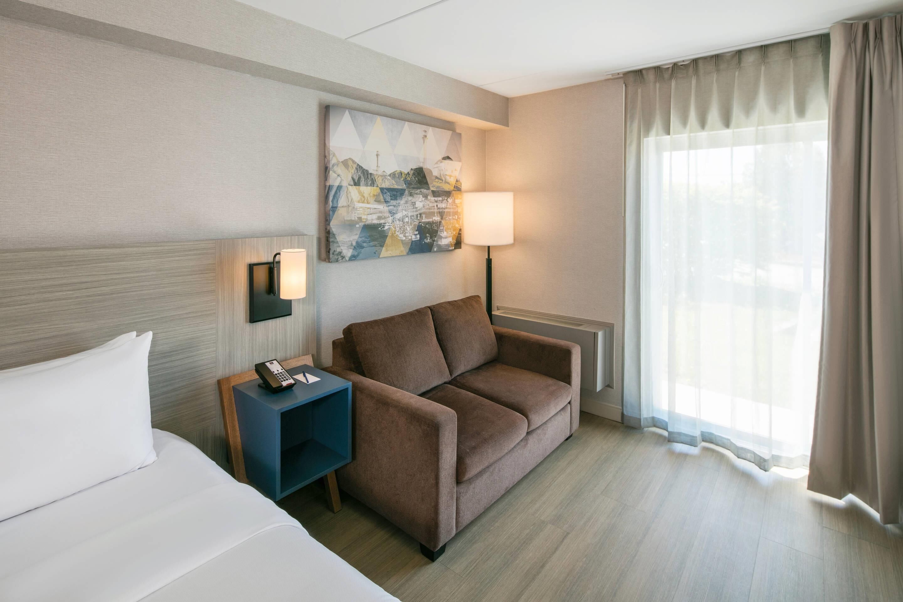 Ein Hotelzimmer mit einem Bett, einem Sofa, einem Schreibtisch mit Lampe, einem Bild an der Wand und einem Fenster mit Vorhängen.