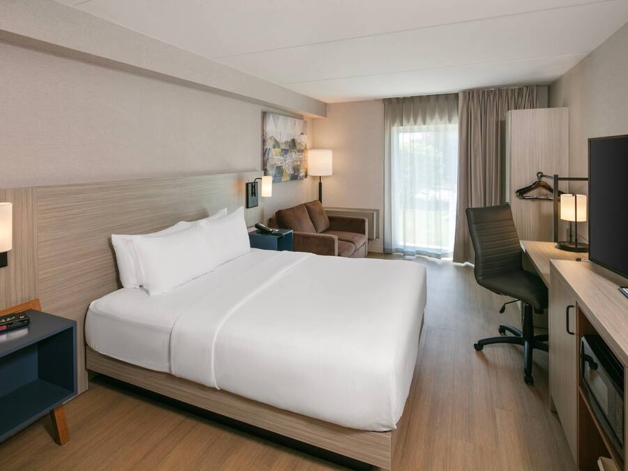 A hotel room with a bed, bedside table, lamp, chair, desk with a TV, and a window with curtains.