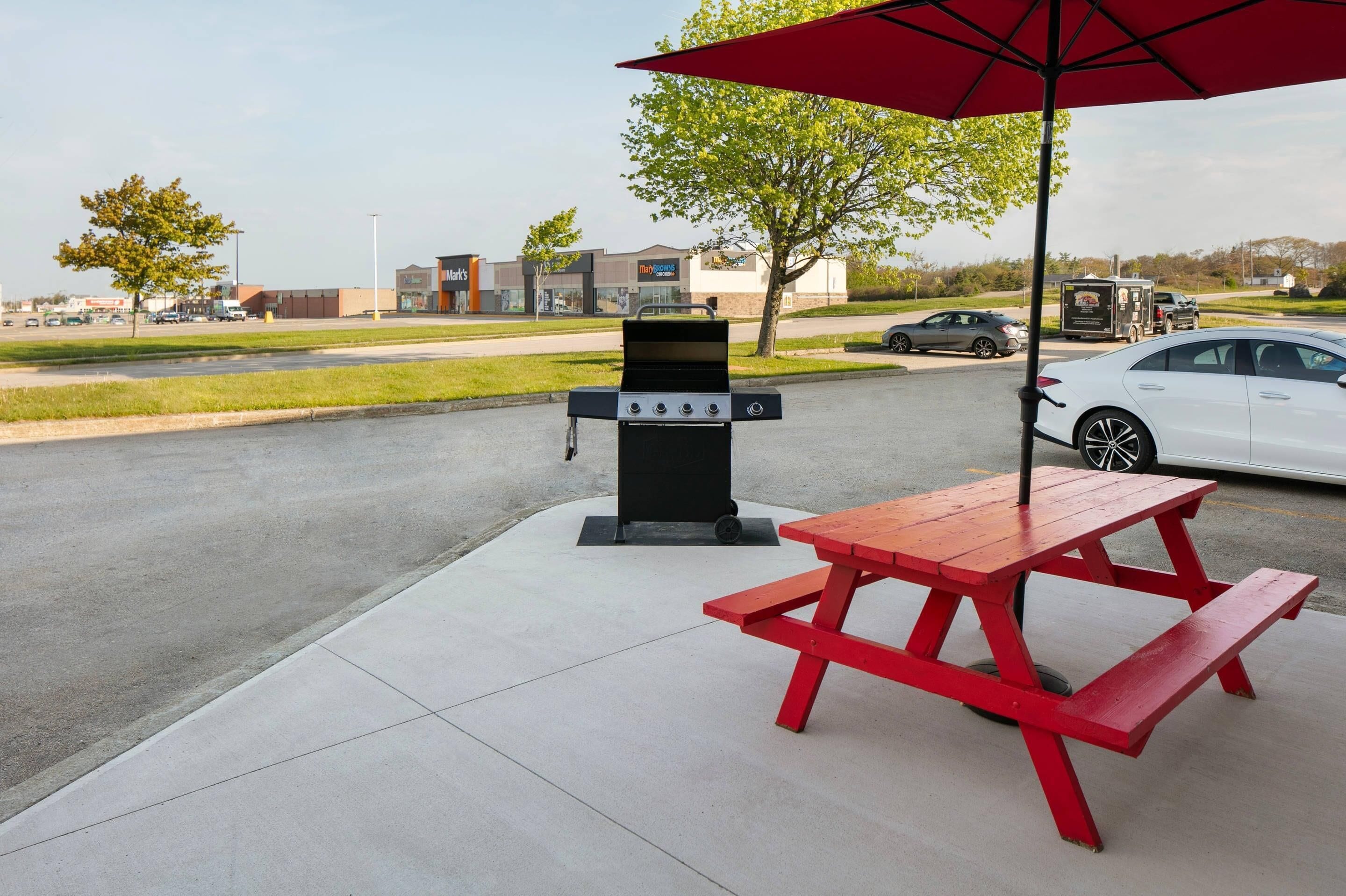 A picnic setup with a grill and table under a red umbrella, located in a parking lot with a food court in the background.