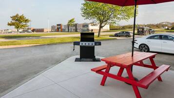 A picnic setup with a grill and table under a red umbrella, located in a parking lot with a food court in the background.