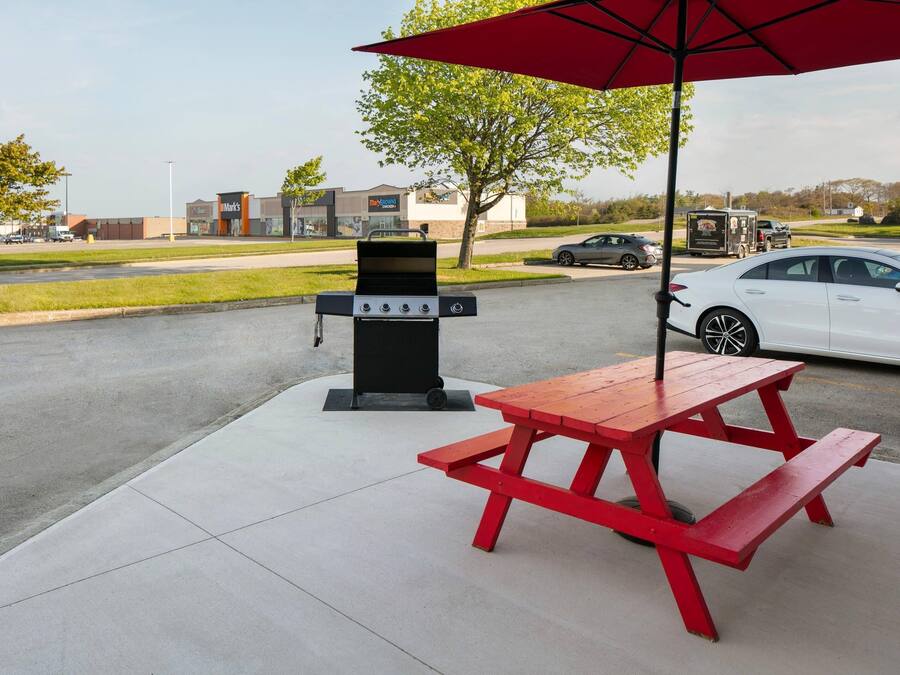 A picnic setup with a grill and table under a red umbrella, located in a parking lot with a food court in the background.
