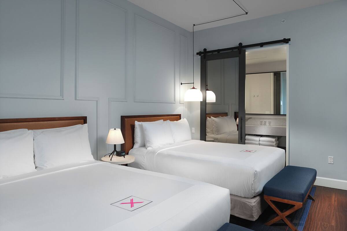 Habitación de hotel con dos camas, una mesita de noche con lámpara, un banco, un espejo y piso de madera.