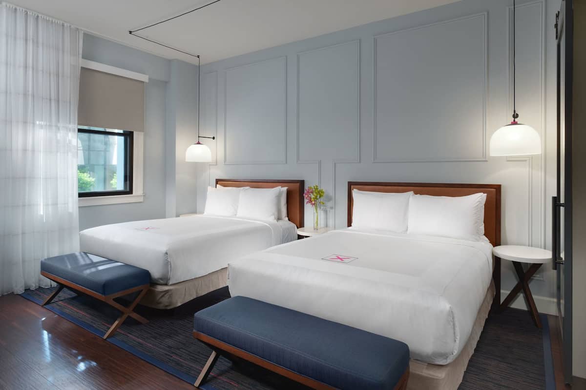 Una habitación de hotel con dos camas, un banco, una mesita y una ventana con cortinas.