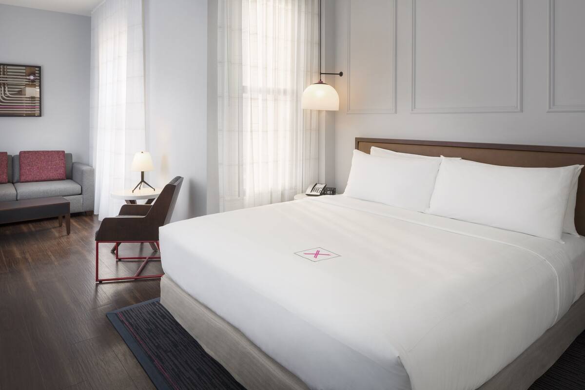 Una habitación de hotel con una cama grande, un escritorio con lámpara, un sofá y una ventana con cortinas traslúcidas.