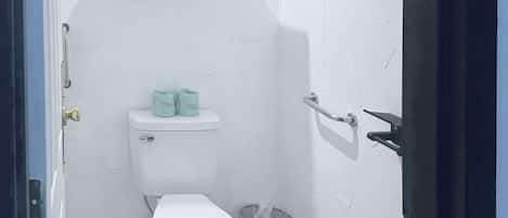 A small, compact bathroom with a toilet, a trash bin, and a shelf above the toilet holding toilet paper.
