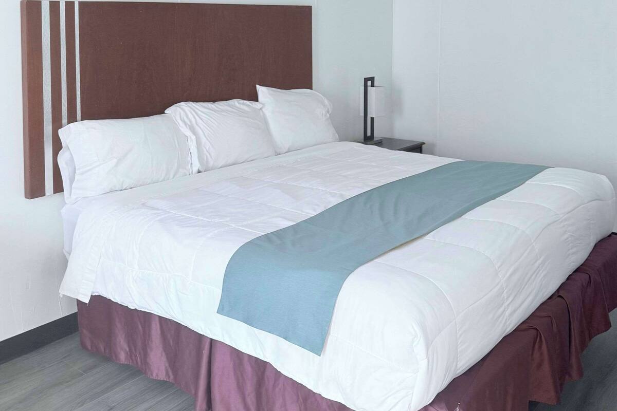 Ein ordentlich bezogenes Bett mit weißer und blauer Tagesdecke, einem Holzkopfende und einem dunklen Nachttisch mit Lampe.