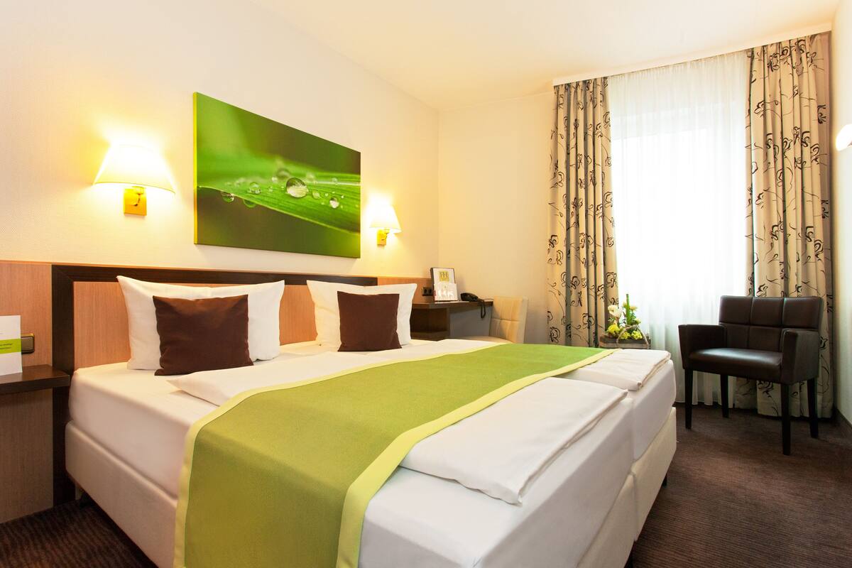 A hotel room with a large bed, two bedside lamps, a green blanket, a chair, and a painting on the wall.
