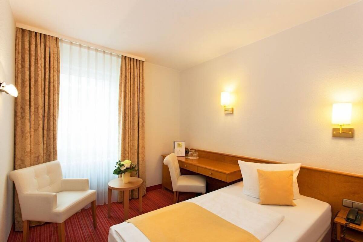 A hotel room with a bed, a desk, a chair, a small table with a vase, and curtains.