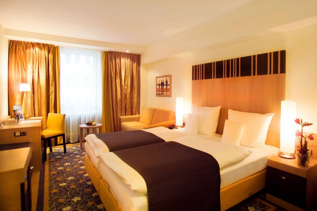 A hotel room with two beds, a desk, a chair, a small table with fruit, and a painting on the wall.