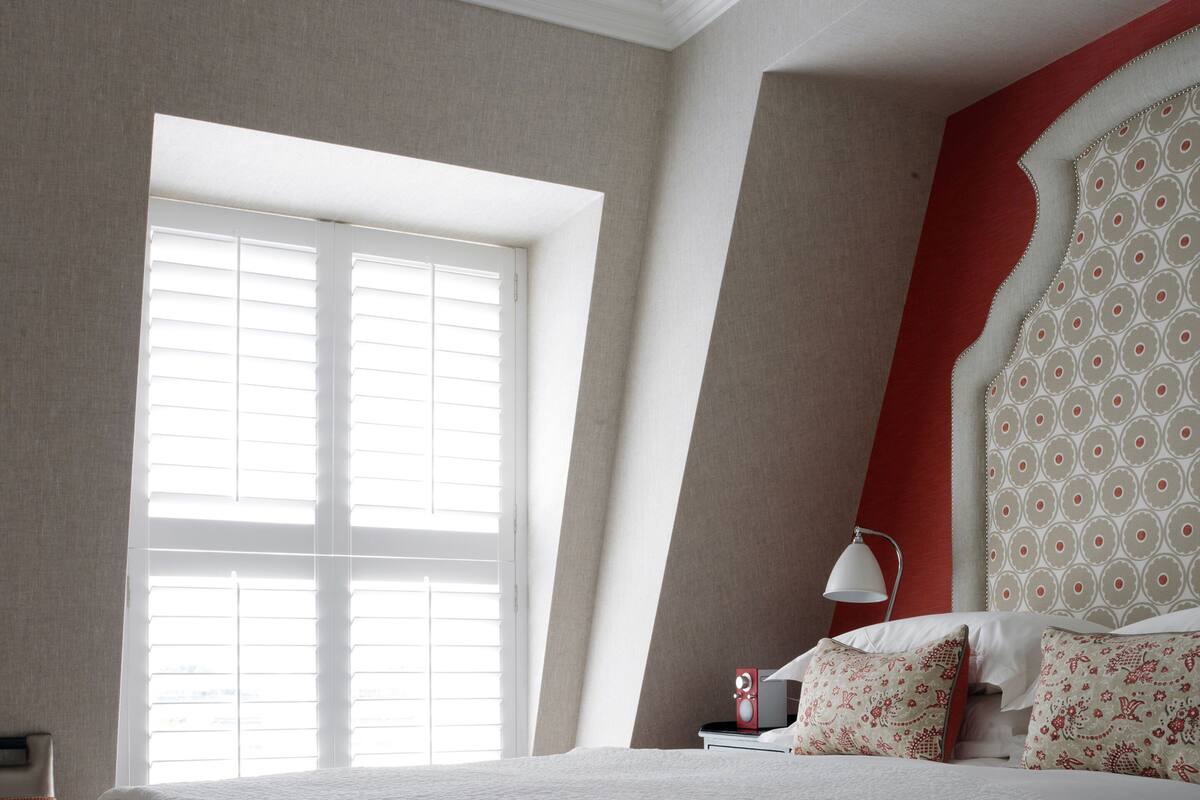 A bedroom with a large window, a bed with white bedding, a red accent wall, and a patterned headboard.