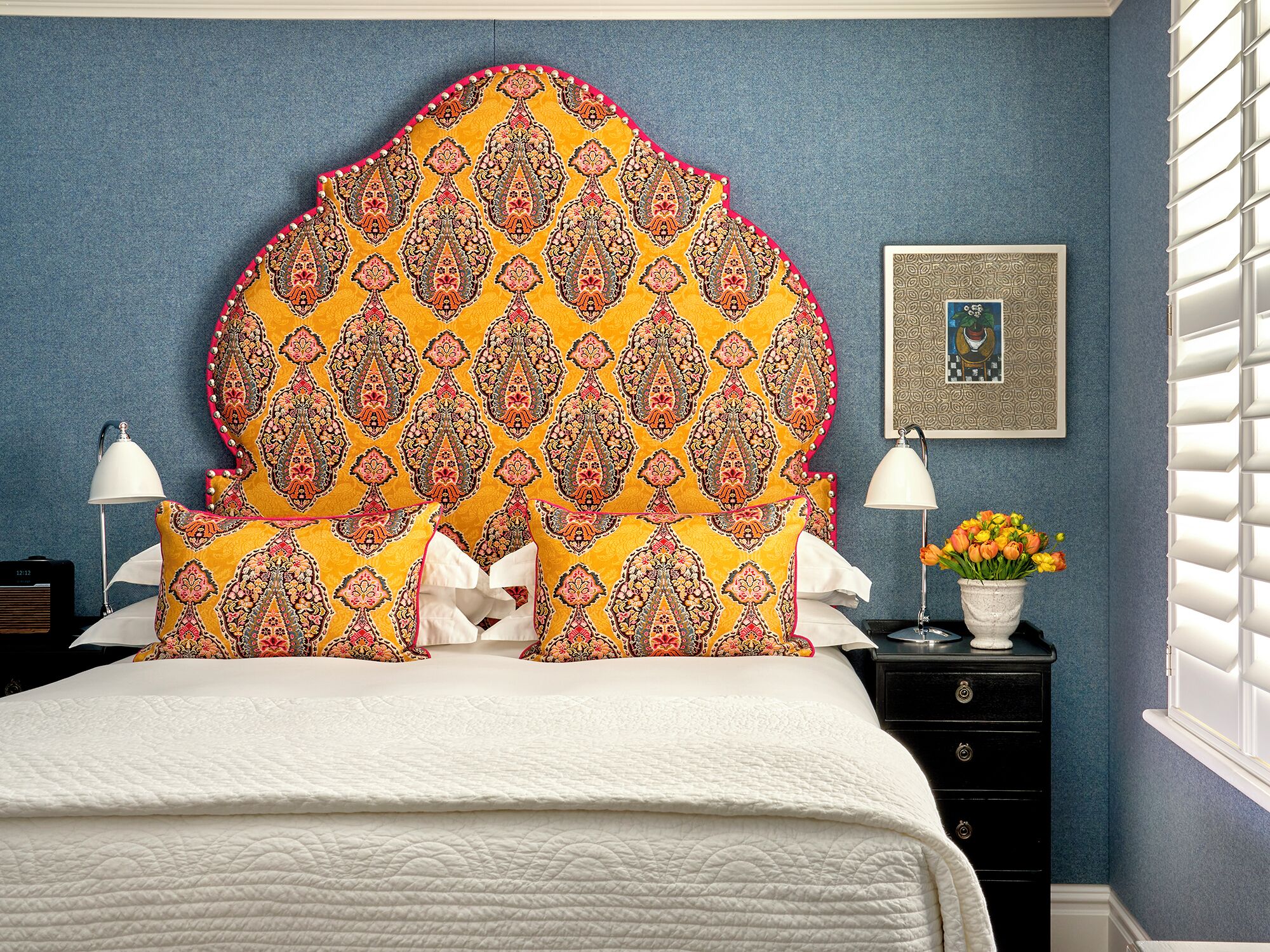 A bed with a white quilt, a decorative headboard with a paisley pattern, two bedside lamps, a nightstand with a vase of flowers, and a framed picture on the wall.