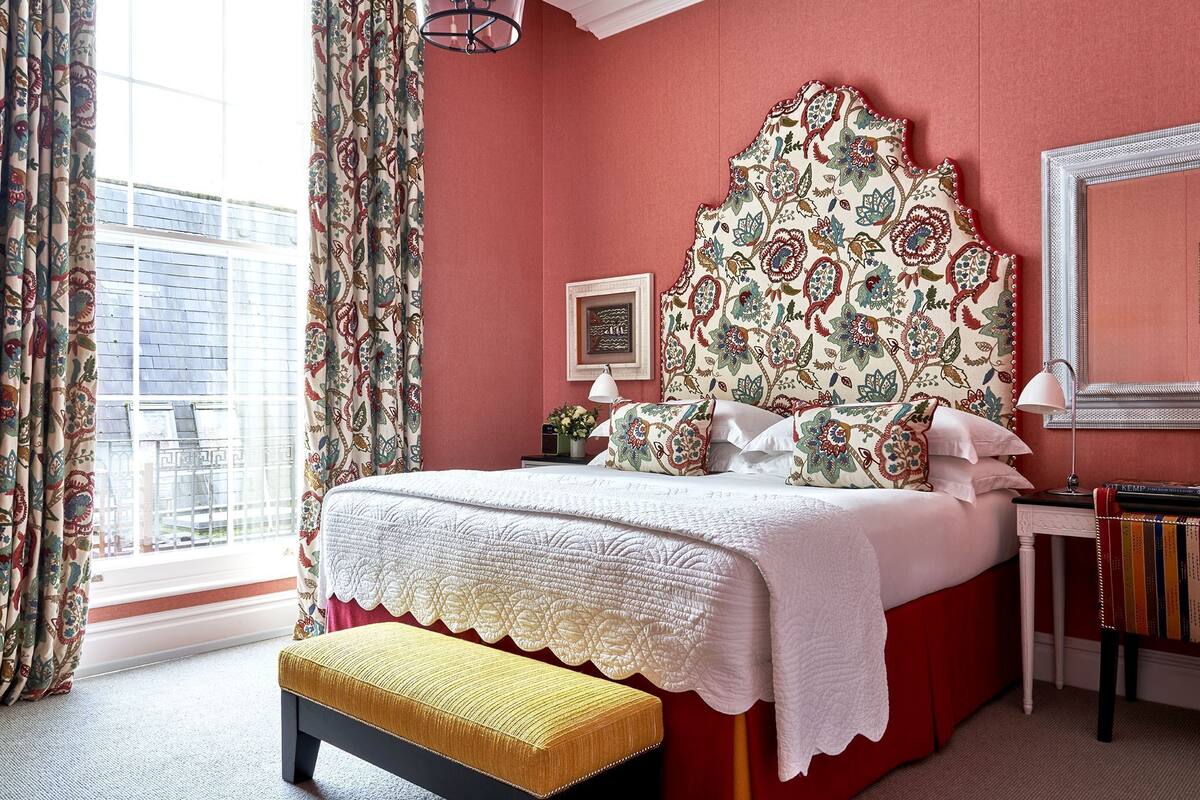 A bedroom with a red wall, a patterned headboard, a white bedspread, a bench, a nightstand with a lamp, and a framed picture.