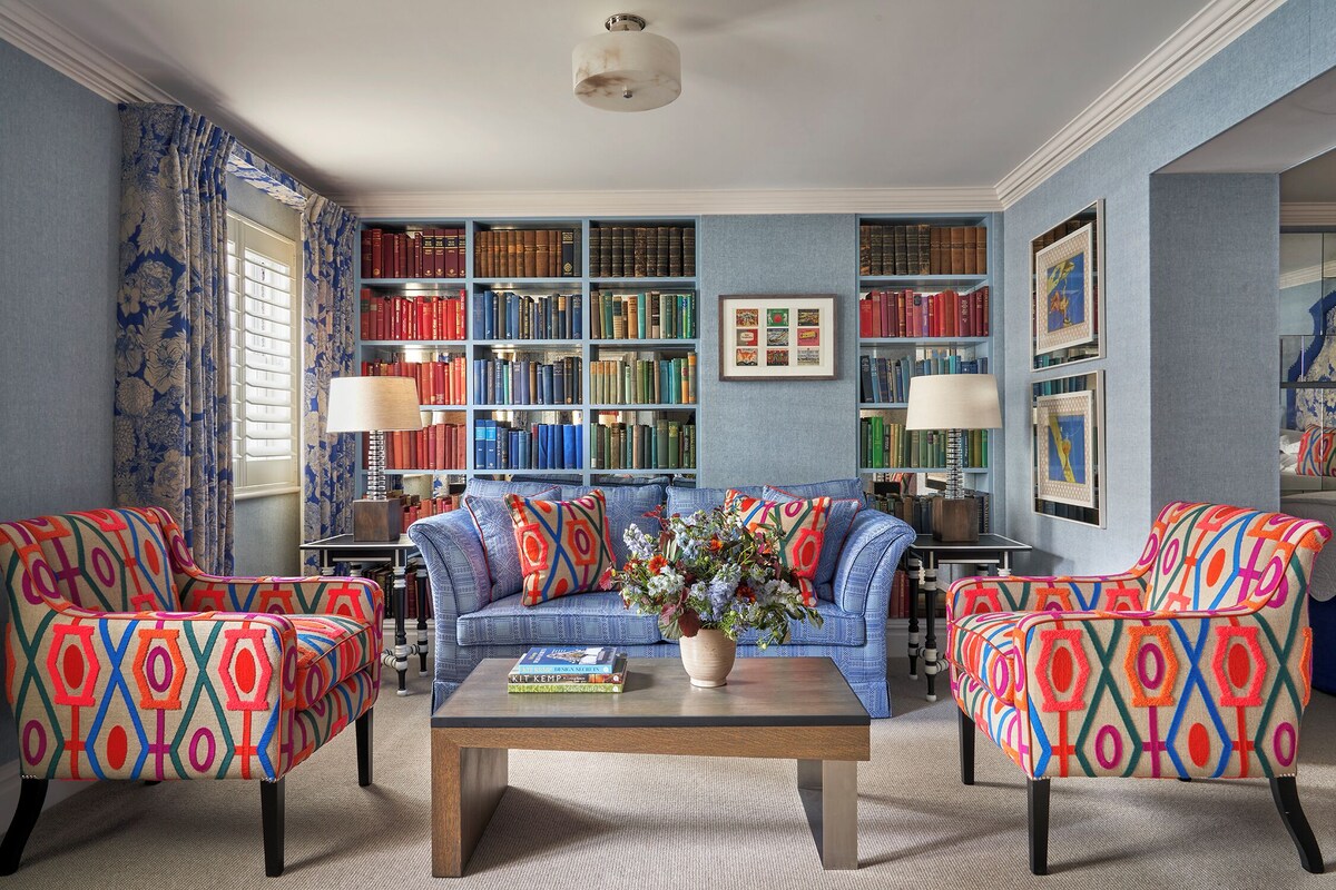 Une pièce avec un canapé bleu, deux fauteuils à motifs, une table basse avec des livres, une bibliothèque et une fenêtre avec des rideaux.