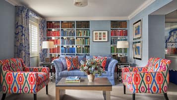 A room with a blue sofa, two patterned armchairs, a coffee table with books, a bookshelf, and a window with curtains.
